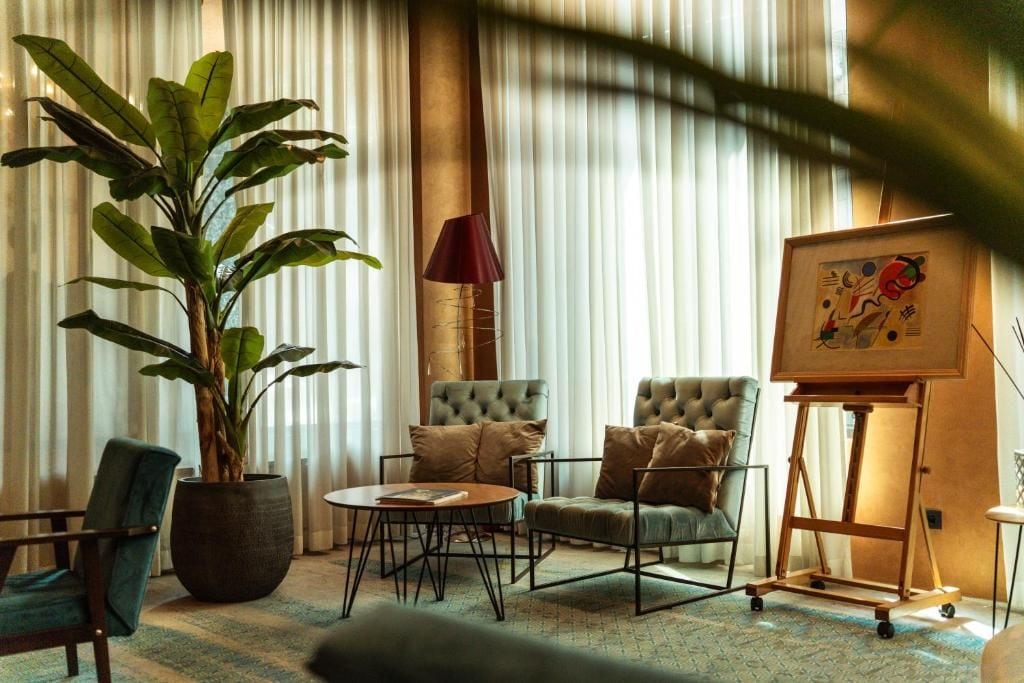 A room with a large potted plant, two tufted armchairs, a round coffee table, a lamp, and a framed artwork on a stand.