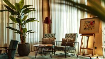 Una habitación con una planta grande en maceta, dos sillones con reposabrazos capitonados, una mesa de centro redonda, una lámpara y una obra de arte en un caballete.
