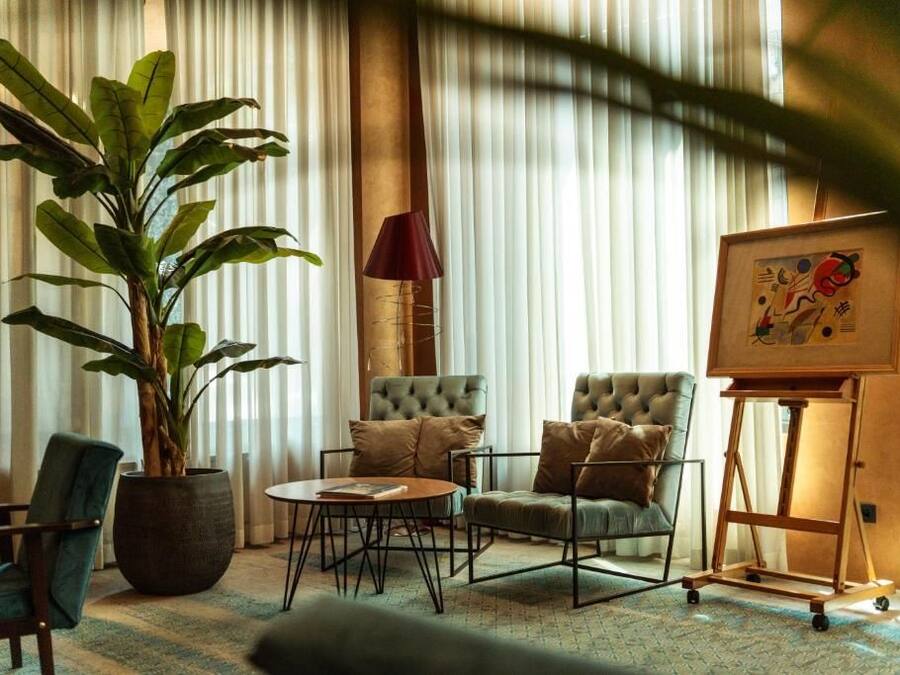 Una habitación con una planta grande en maceta, dos sillones con reposabrazos capitonados, una mesa de centro redonda, una lámpara y una obra de arte en un caballete.