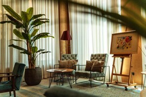 A room with a large potted plant, two tufted armchairs, a round coffee table, a lamp, and a framed artwork on a stand. - Crystal Hotel Belgrade (Belgrade)