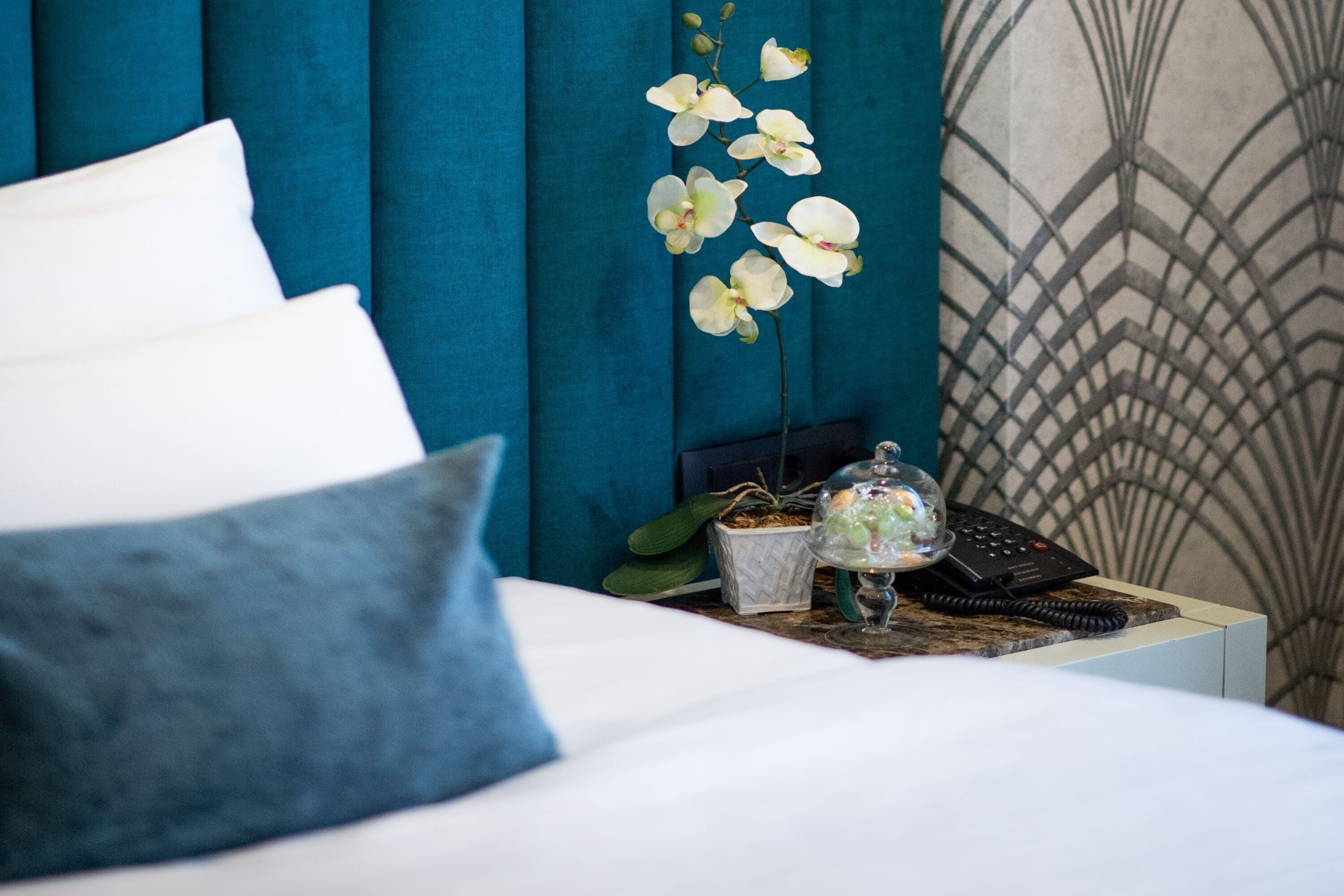 A neatly made bed with white and blue pillows, a small table with a phone and decorative items, and a wall with a geometric pattern.