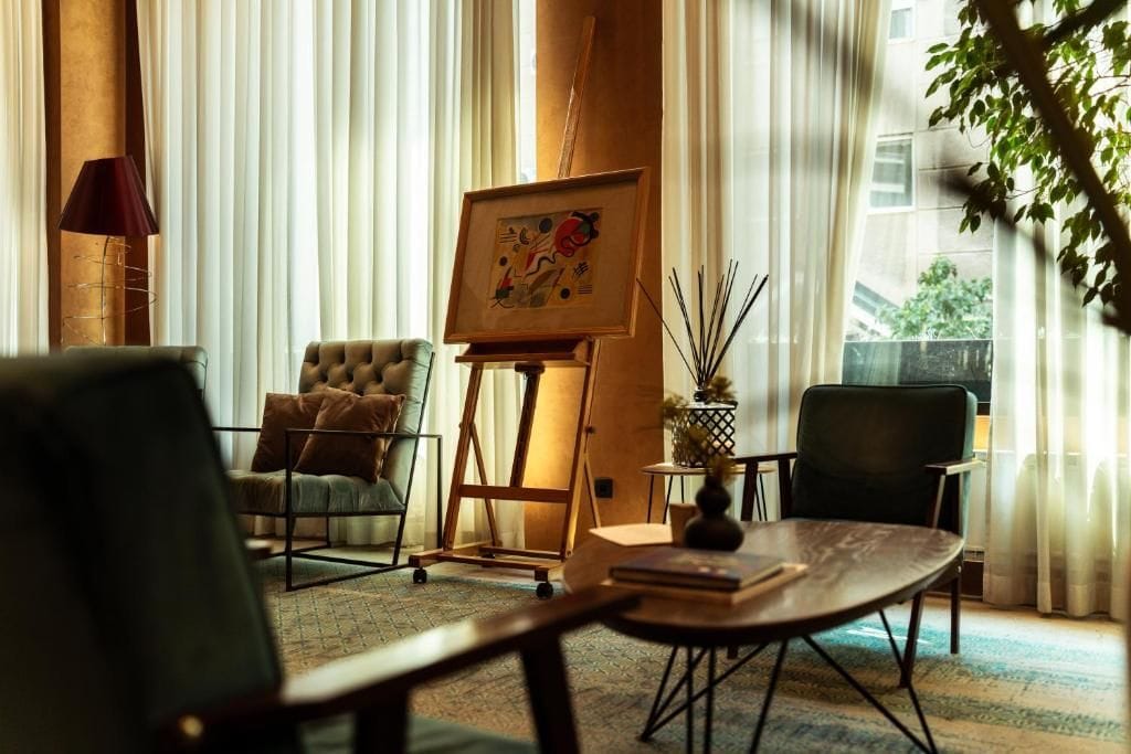 A room with a large window, a painting on easel, a sofa, a chair, and a table with books.
