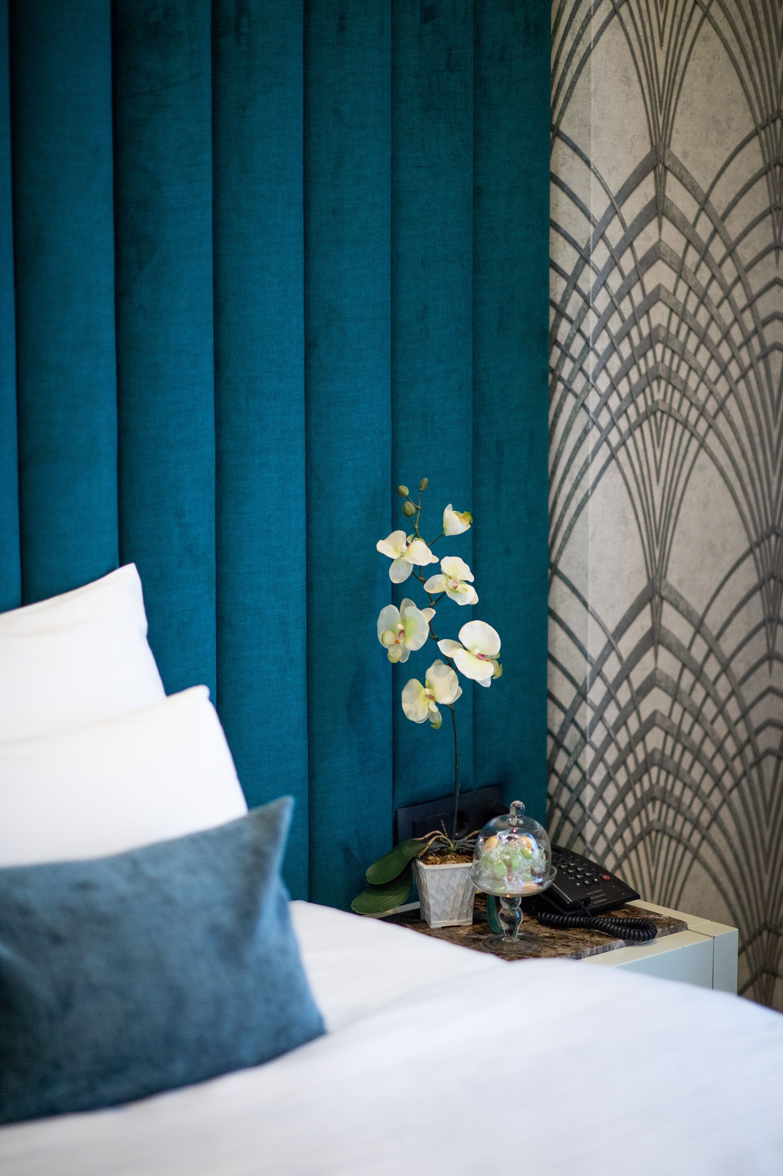Una cama bien hecha con almohadas blancas y azules, una mesita de noche con un florero de orquídeas blancas y una pared con un diseño decorativo.