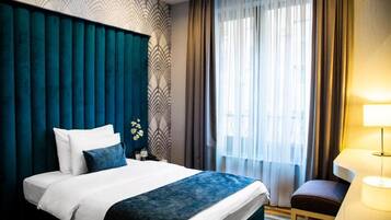 A hotel room with a large bed, a bedside table with a lamp, a window with curtains, and a patterned wall.