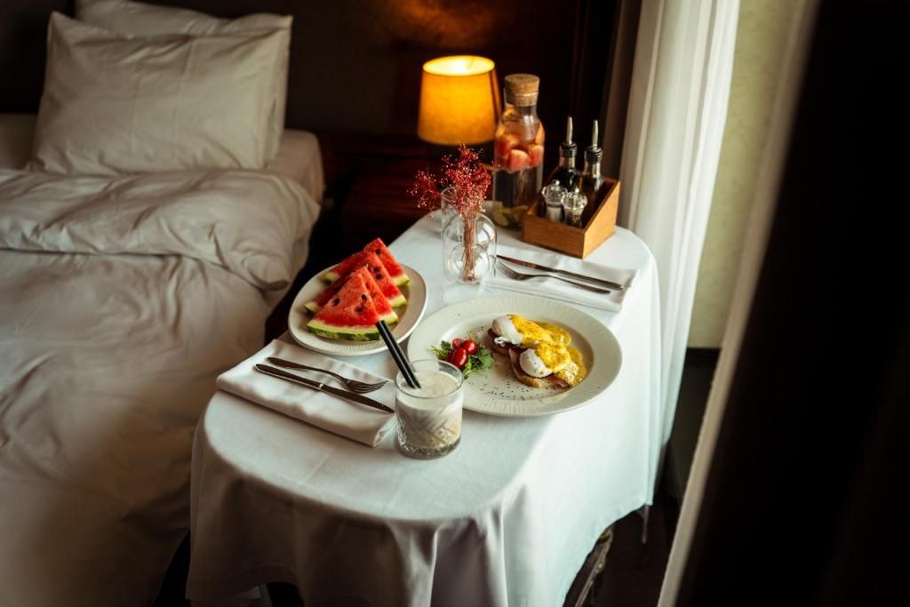 A neatly made breakfast table with plates of food, a vase with dried flowers, and a lamp on a bedside table in a hotel room.