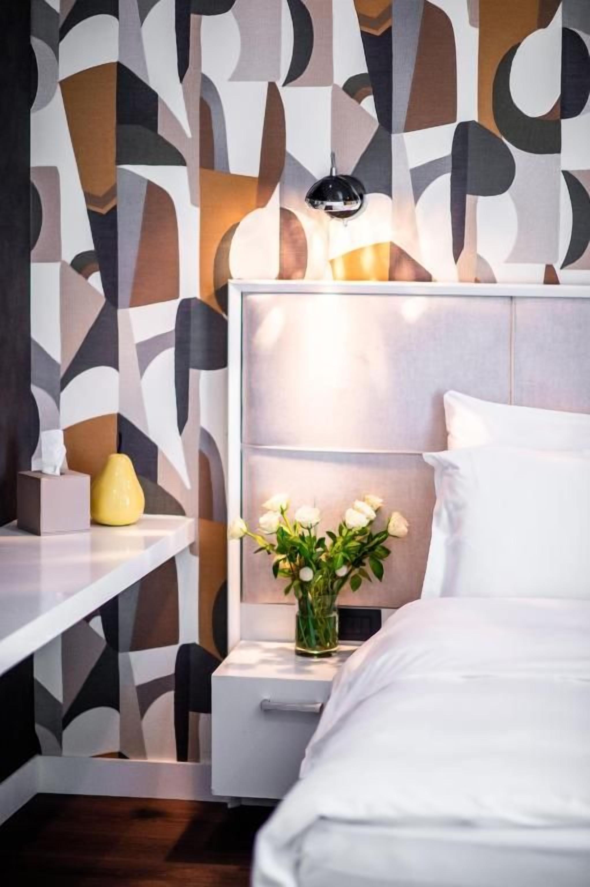 A modern hotel room with a bed, bedside table, and a wall with abstract patterns.