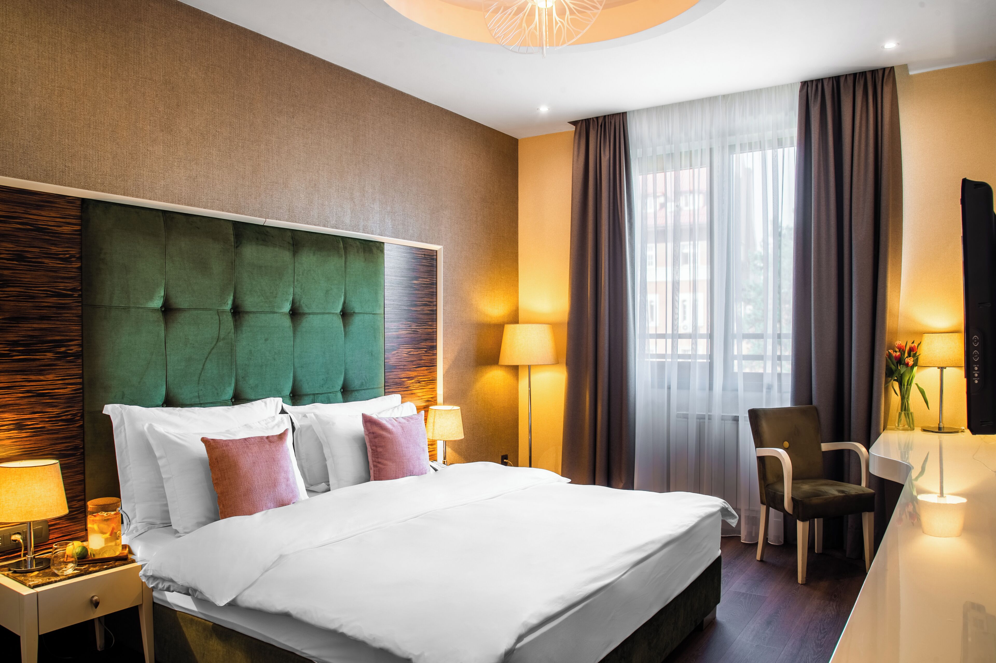 Habitación de hotel con una cama grande, un escritorio con silla, una lámpara y vistas al exterior a través de la ventana.