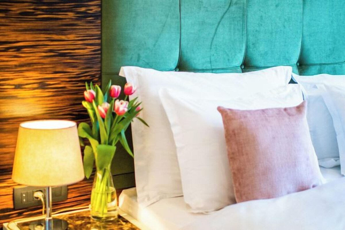 A bed with white and pink pillows, a vase with flowers, and a lamp on a bedside table.