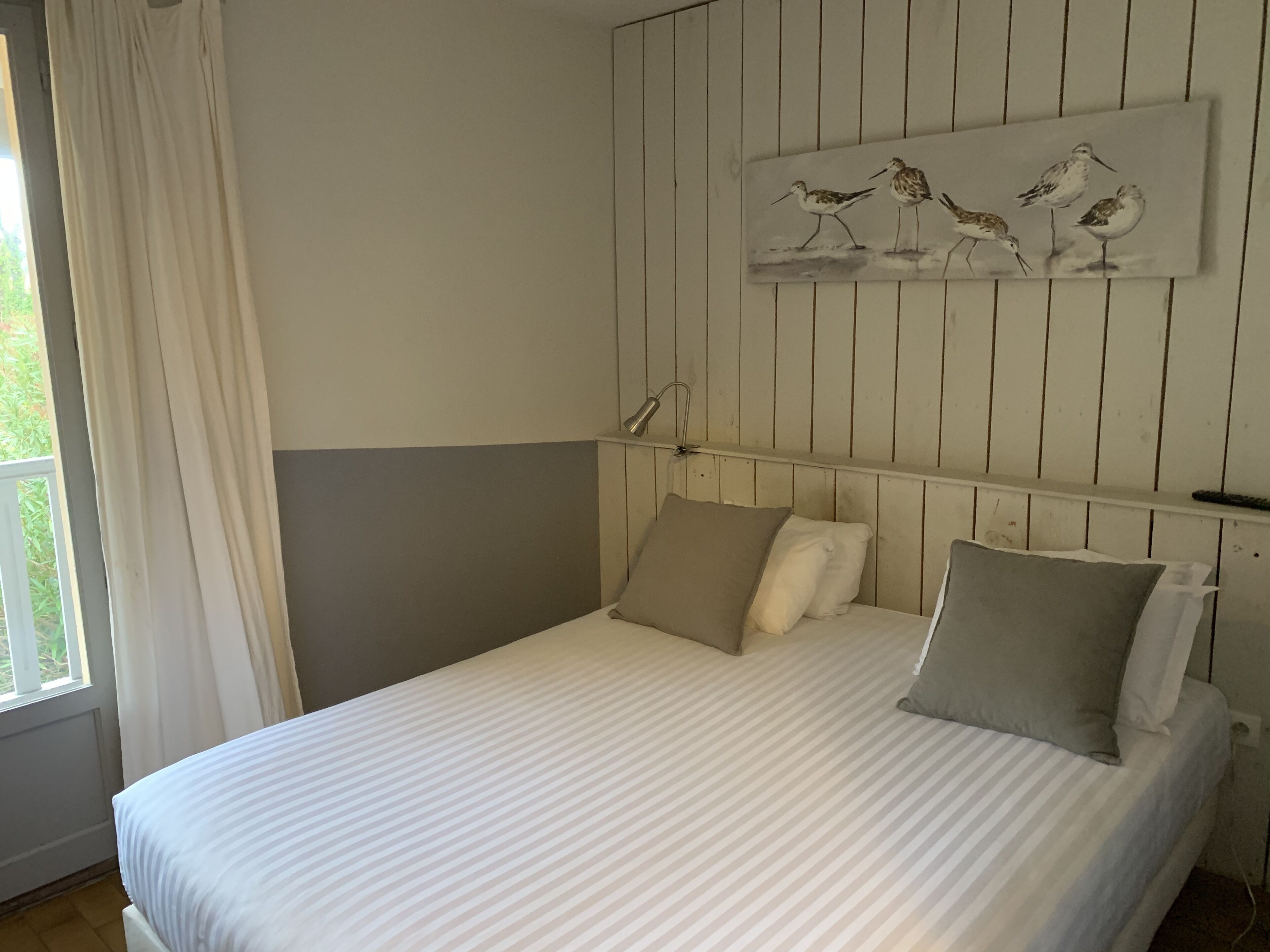 Een slaapkamer met een bed, kussens, een nachtkastje, een lamp en een schilderij van vogels aan de muur.