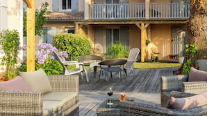 Una terraza de madera con mobiliario de mimbre, incluyendo sofás y sillones, y una mesa de vidrio con una copa de vino y una bebida.