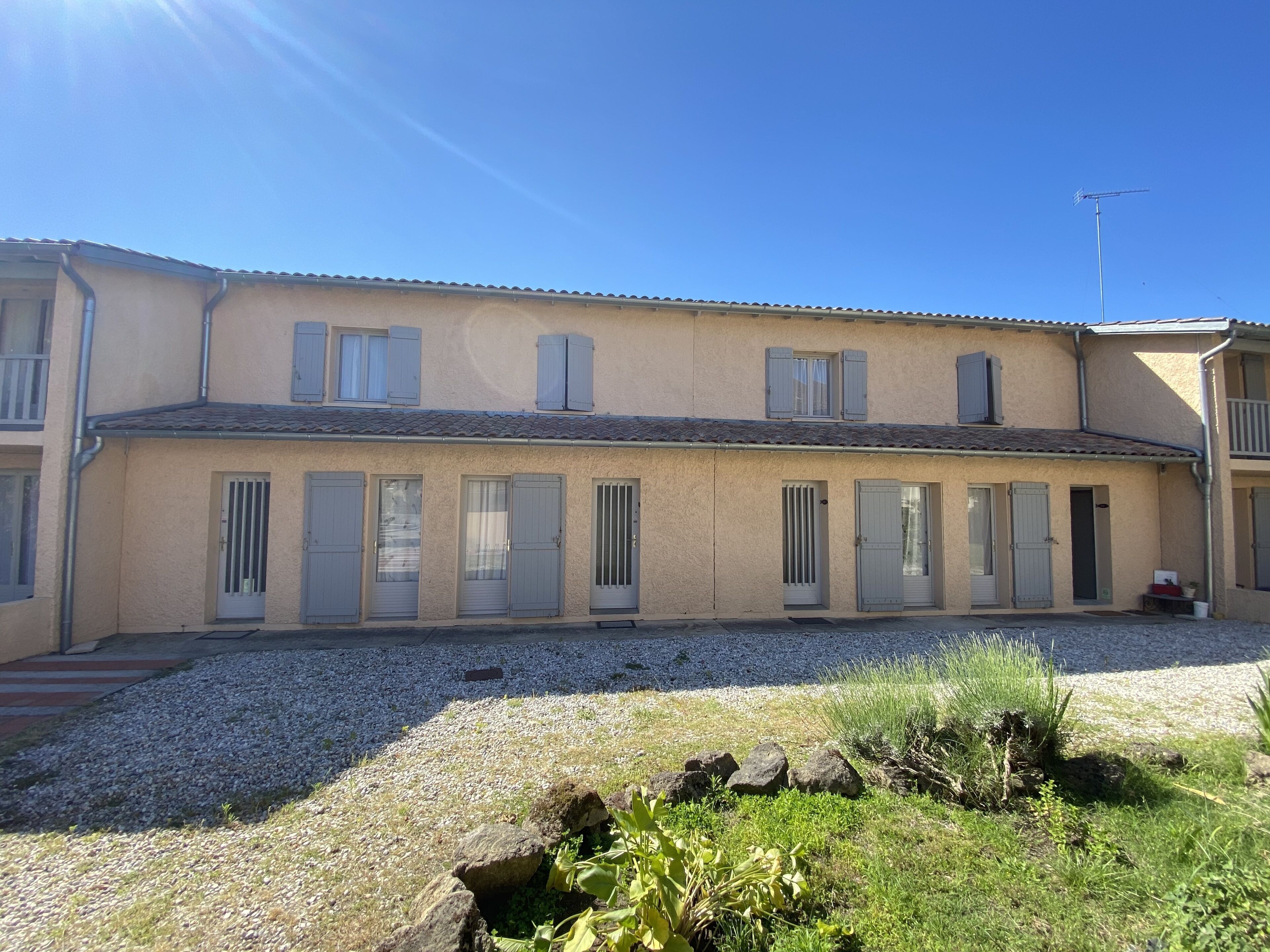 A two-story building with a tiled roof, multiple windows with shutters, and a gravel driveway.