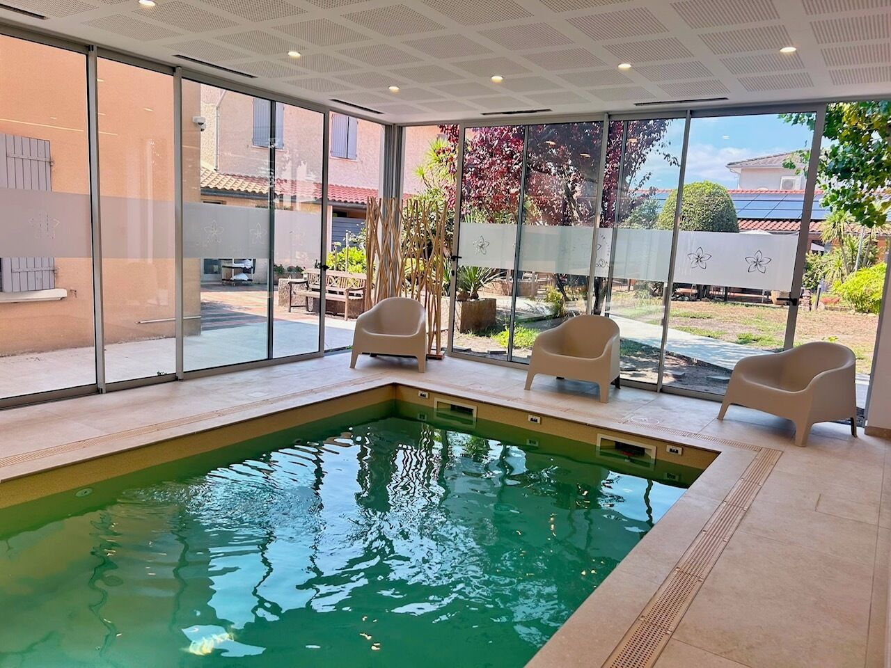 Uma piscina interior com paredes de vidro transparente, três cadeiras bege e vista para o exterior.