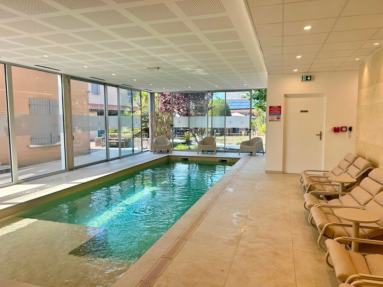 An indoor swimming pool with clear glass walls, a tiled floor, and lounge chairs along the side.