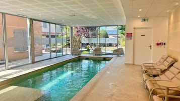 Una piscina interior con paredes de vidrio transparente, piso de baldosas y sillas de descanso a lo largo.