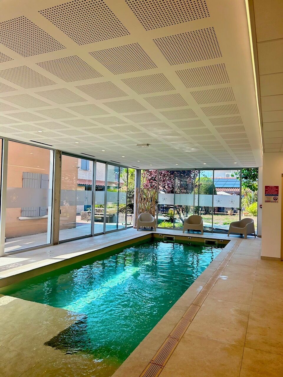 An indoor swimming pool with clear glass walls, a tiled floor, and a ceiling with a geometric pattern.