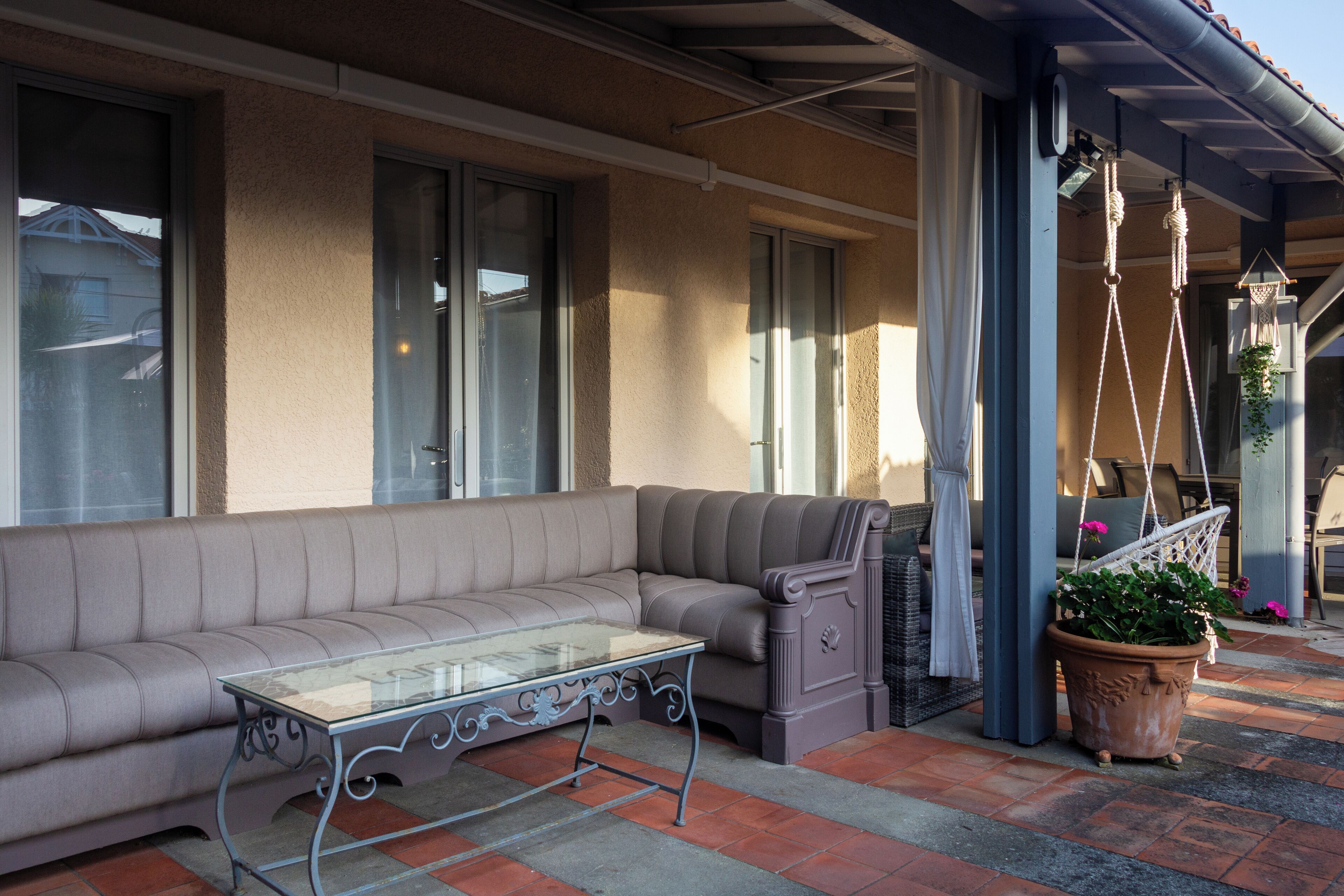 Een overdekt terras met een bank, een tafel met glazen blad en een potplant.