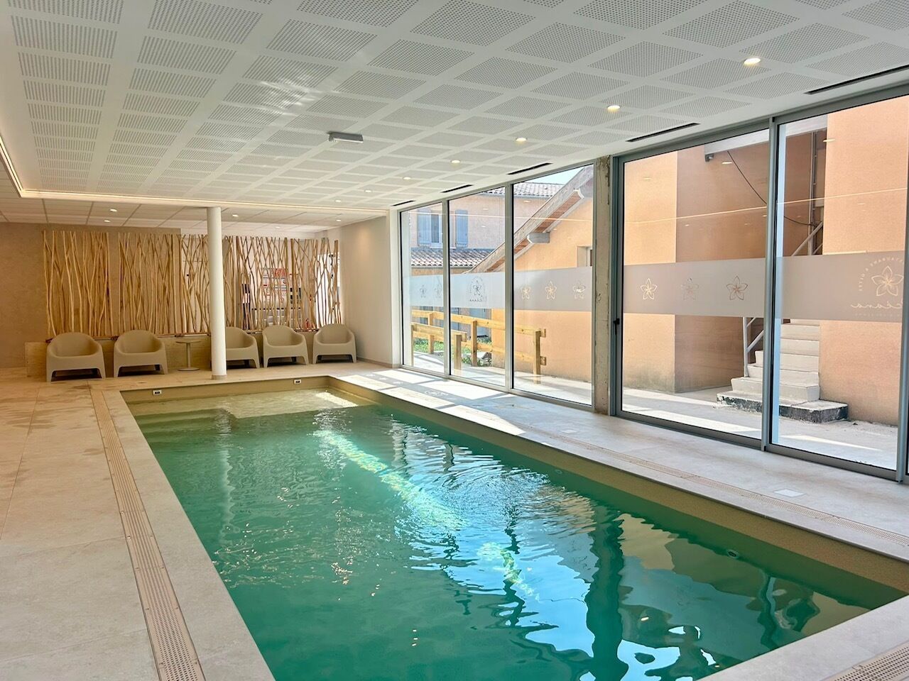 Una piscina interior con paredes de vidrio transparente, techo con diseño de cuadrícula y zona de estar con sillas.