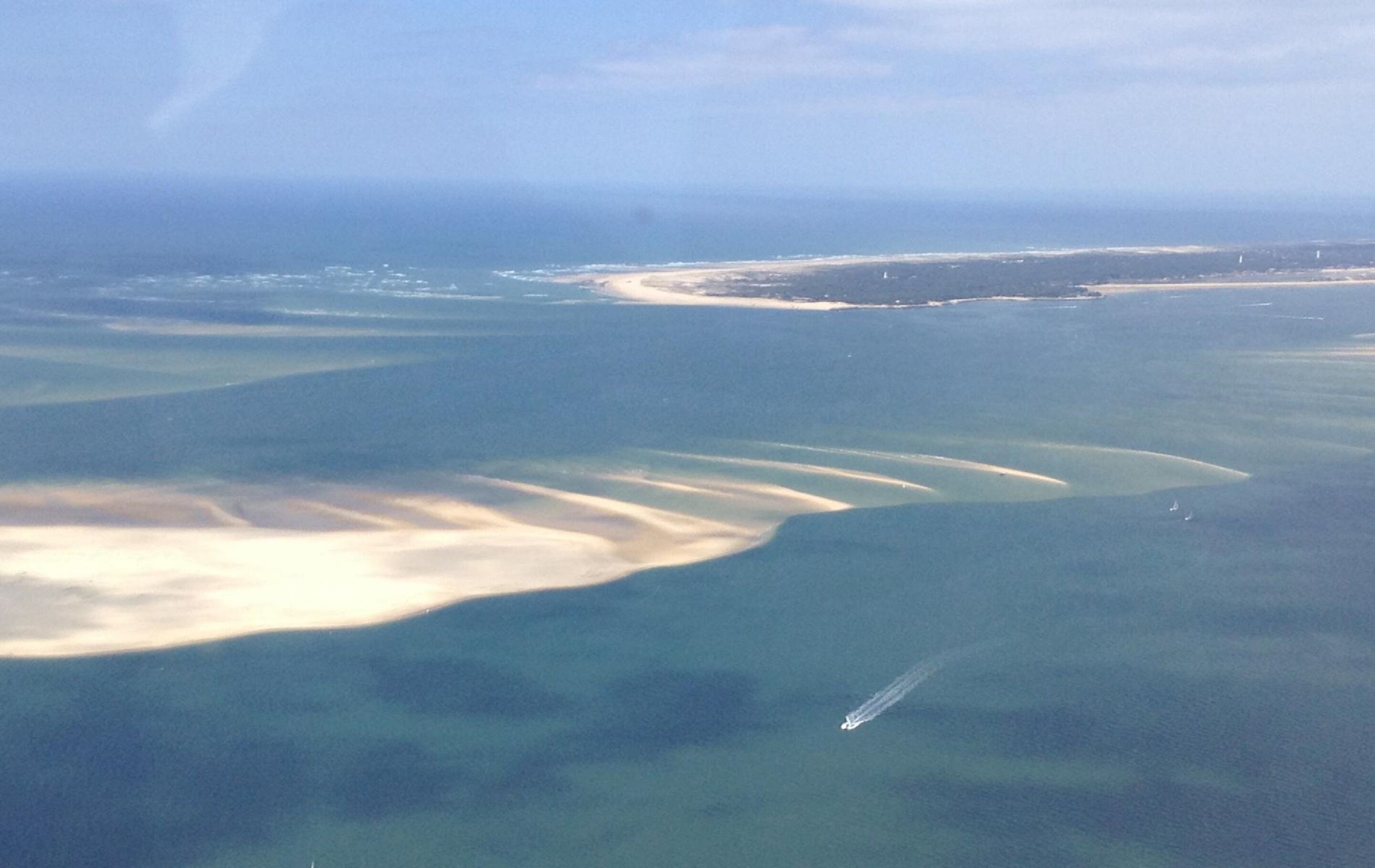 Vue aérienne d’une zone côtière avec une plage de sable, une petite île et un bateau sur l’eau.