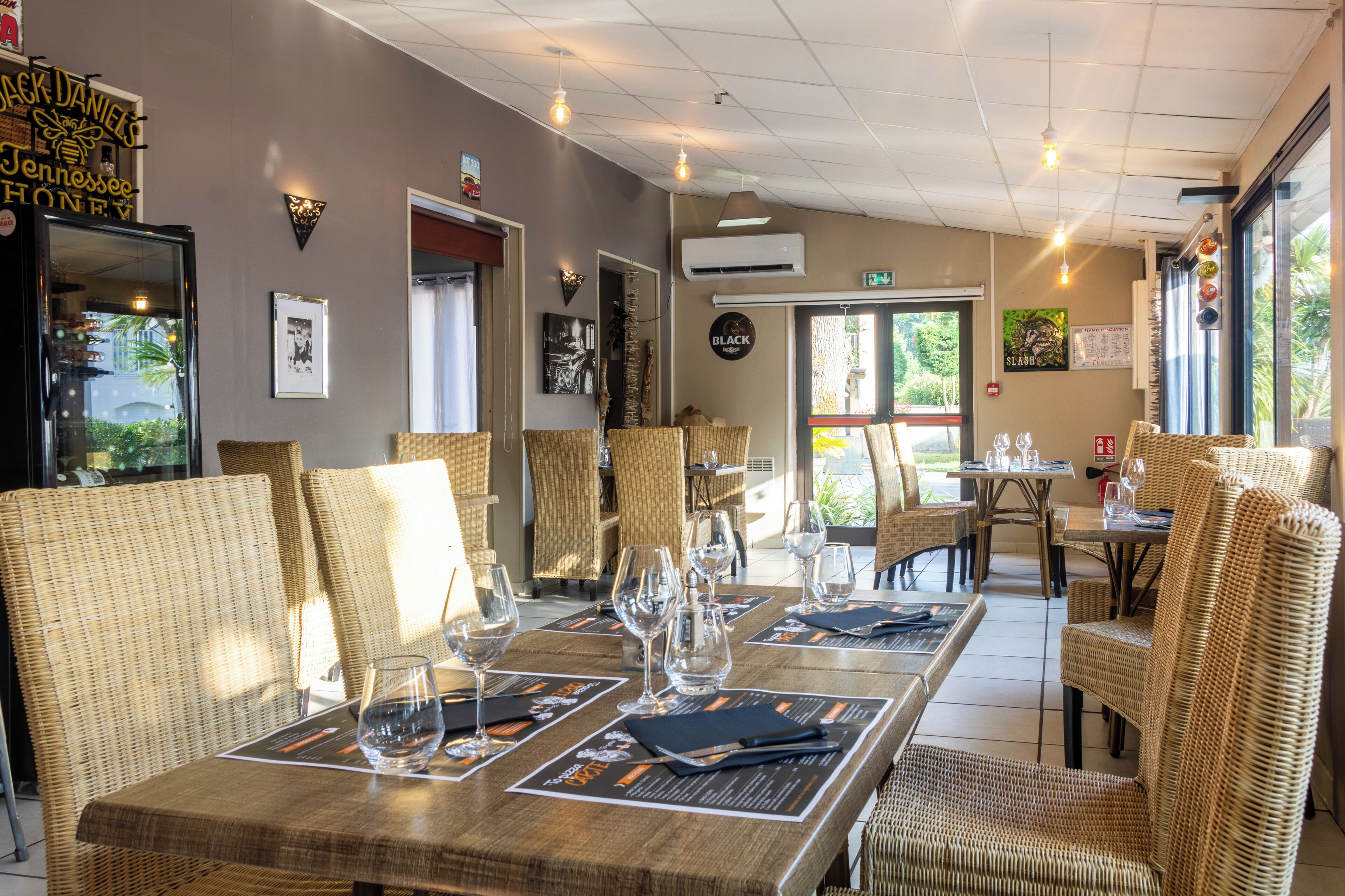 Restaurante com móveis de vime, mesas preparadas com copos de vinho e cardápios, e vista para o exterior.