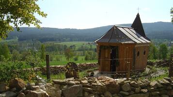 A small wooden cabin with a sloped roof and a cross on top, surrounded by a stone wall and lush greenery.