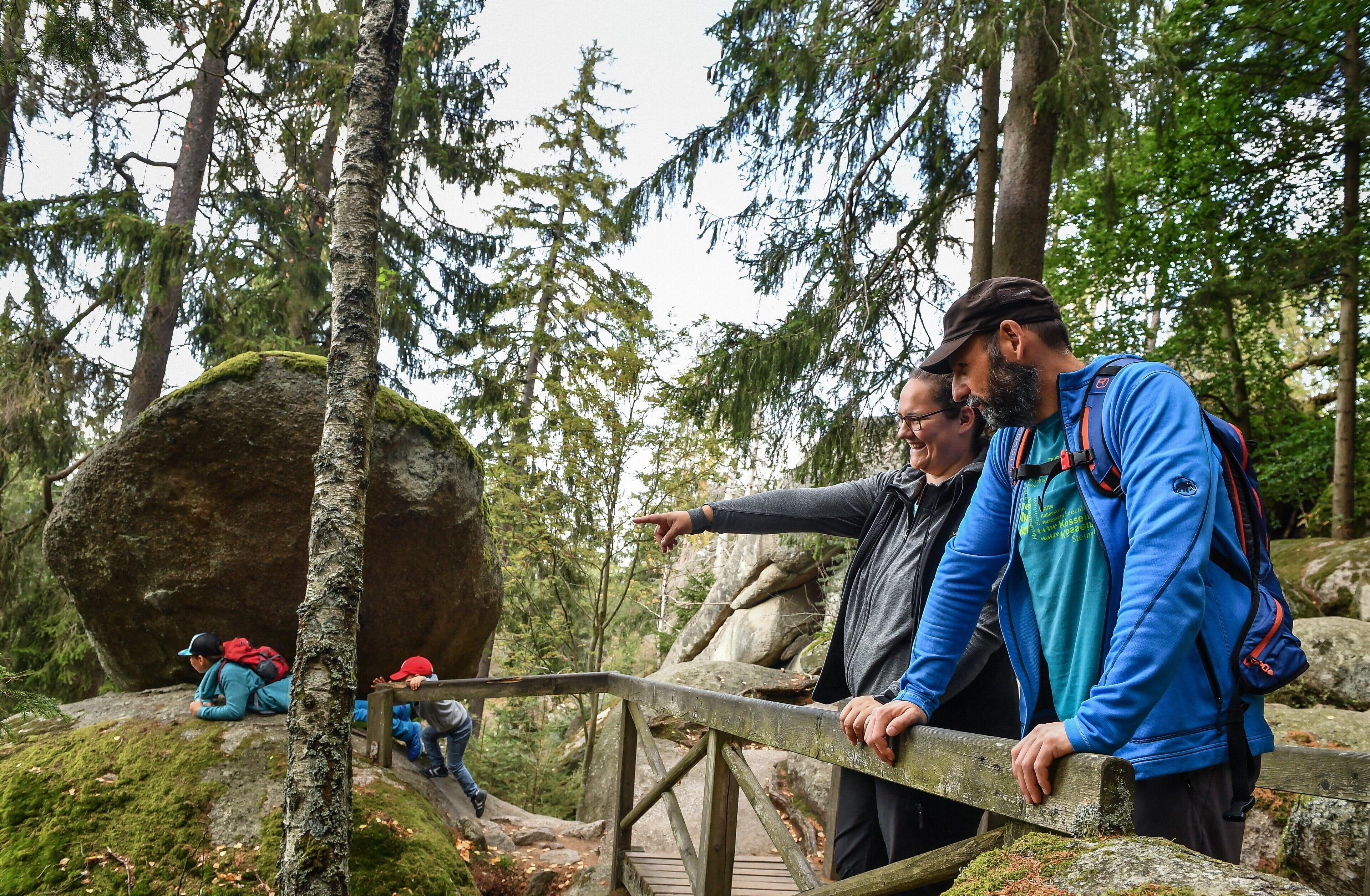Eine Holzbrücke in bewaldeter Landschaft, mit Wanderern und einem großen, moosbewachsenen Felsen in der Nähe.
