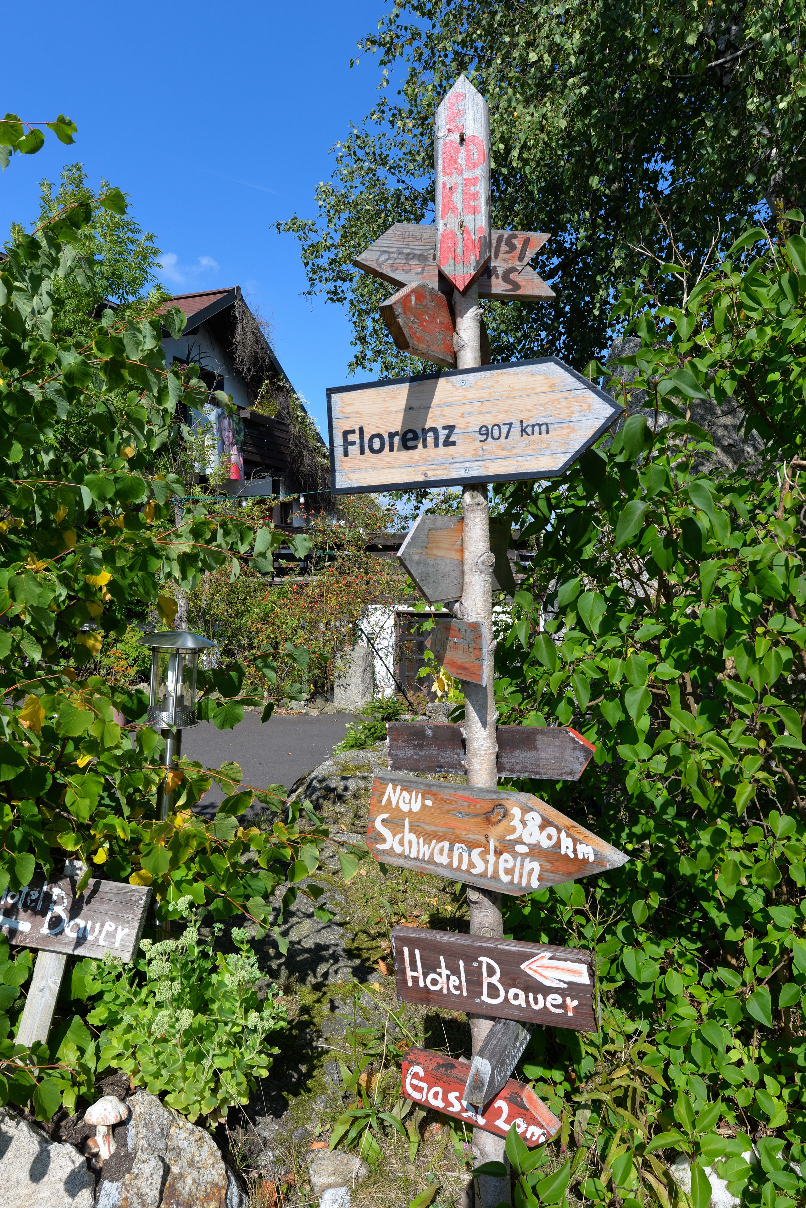 A directional signpost with multiple arrows pointing to various destinations including Florenz, 907 km, Neuschwanzstein, Hotel Bauer, and Gasthof.