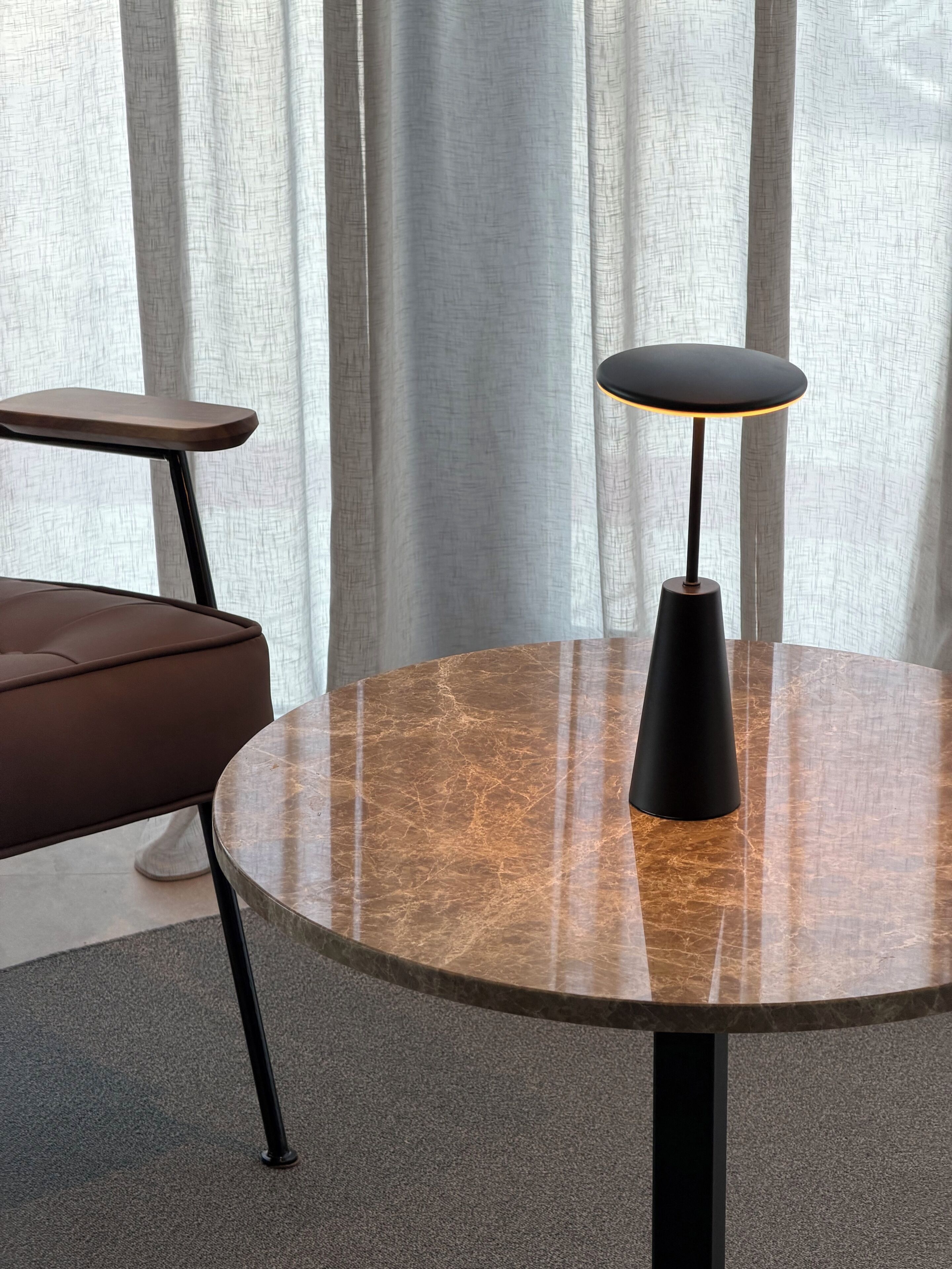 A round table with a marble top, a black lamp, and a brown chair in a room with sheer curtains.
