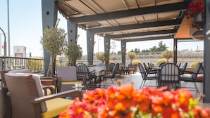 Un patio al aire libre con mobiliario de mimbre, mesas y sillas, rodeado de macetas con plantas y flores.