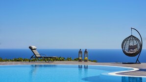 A pool with a clear view of the ocean, a lounge chair, a hanging chair, and two lanterns. - Golden East Hotel (Santorini)