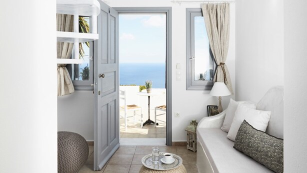 A room with a white interior, a balcony with a view, and a white sofa with cushions. - Golden East Hotel (Santorini)