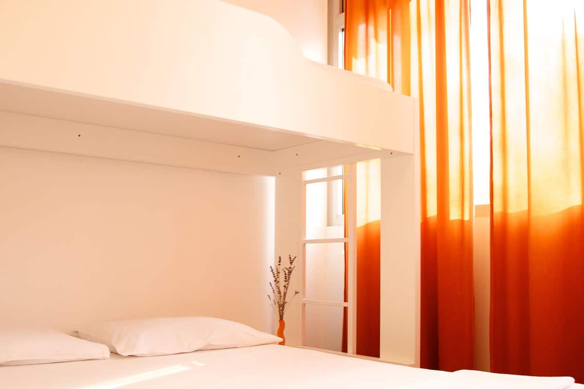 Ein Etagenbett mit weißem Kopfteil, ein Nachttisch mit Blumenvase und orangefarbene Vorhänge.