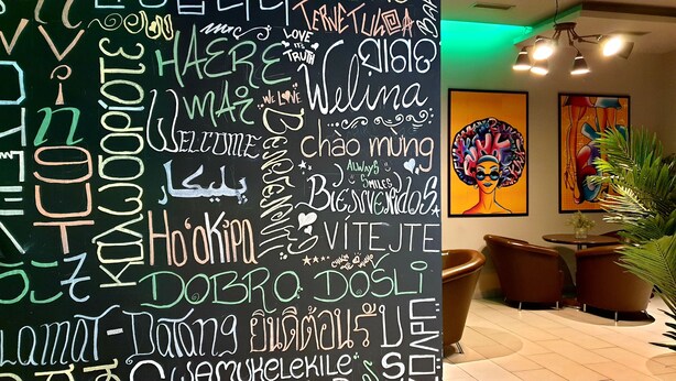 Una pared con diversas frases y palabras en múltiples idiomas, una obra de arte moderno, y un área de descanso con sillas y una mesa.