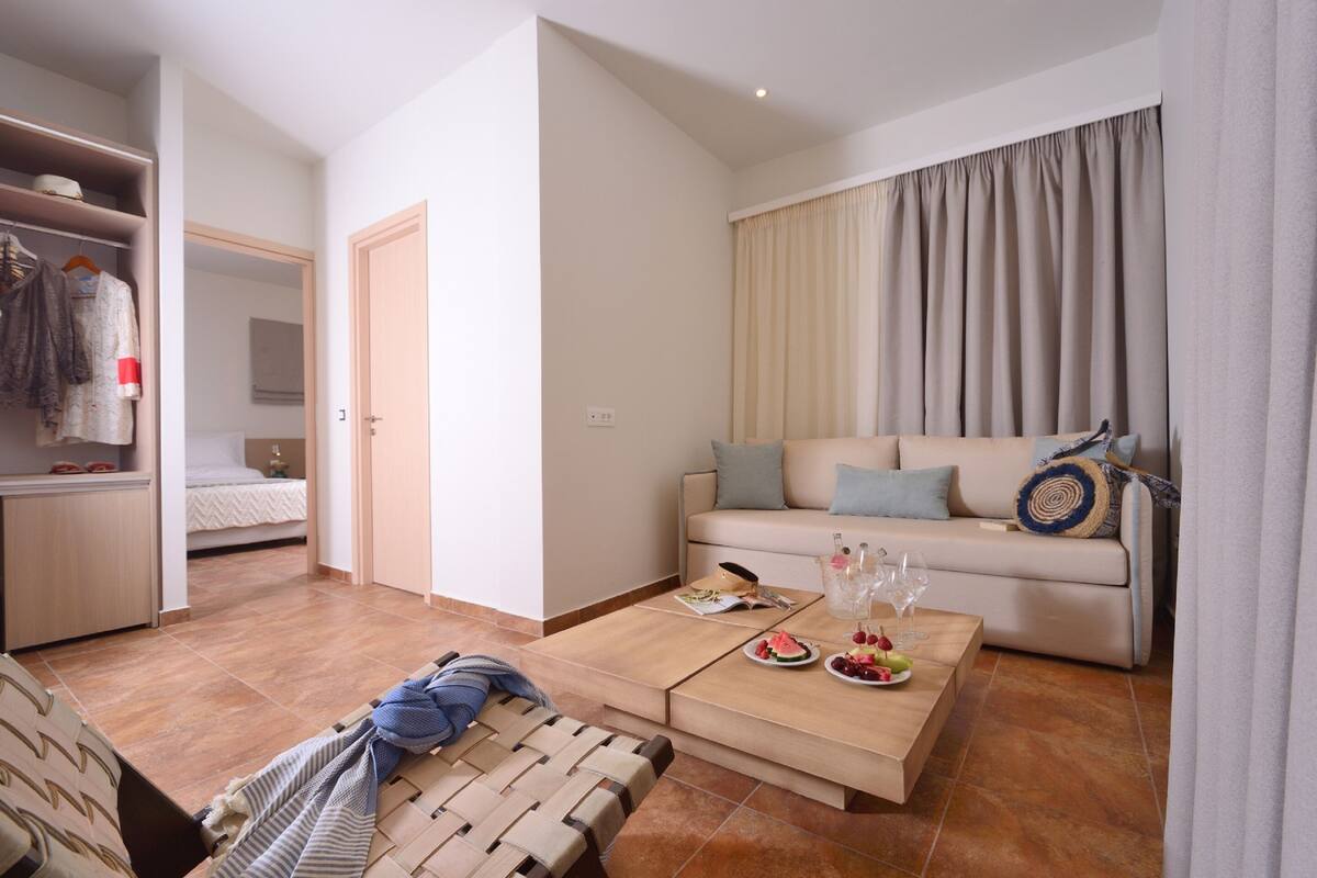 A modern living room with a sofa, a coffee table with food and drinks, a wardrobe, and a bed in the background.
