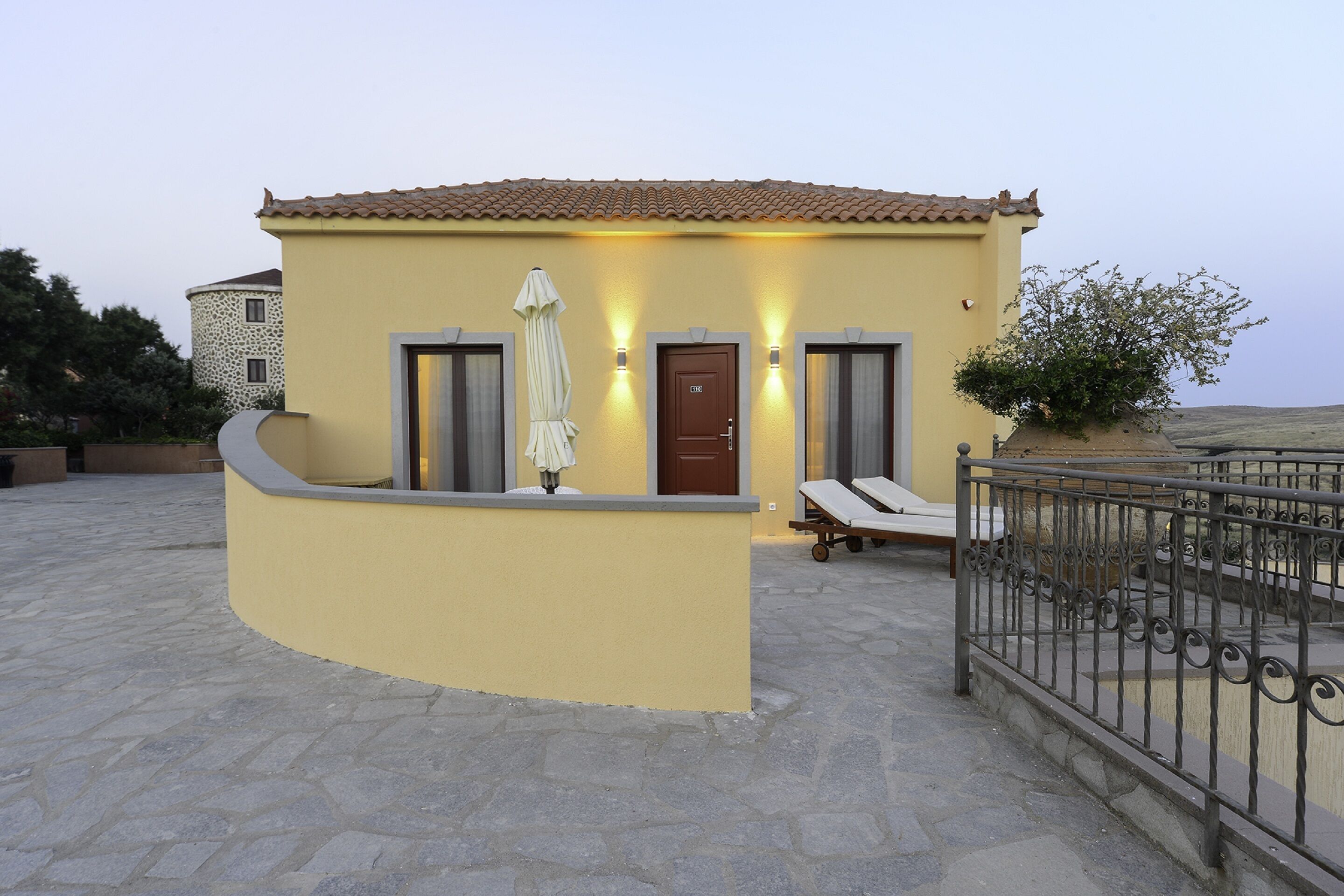 A yellow building with a tiled roof, a door, and two windows with shutters. There is a small garden area with a bench and an umbrella.