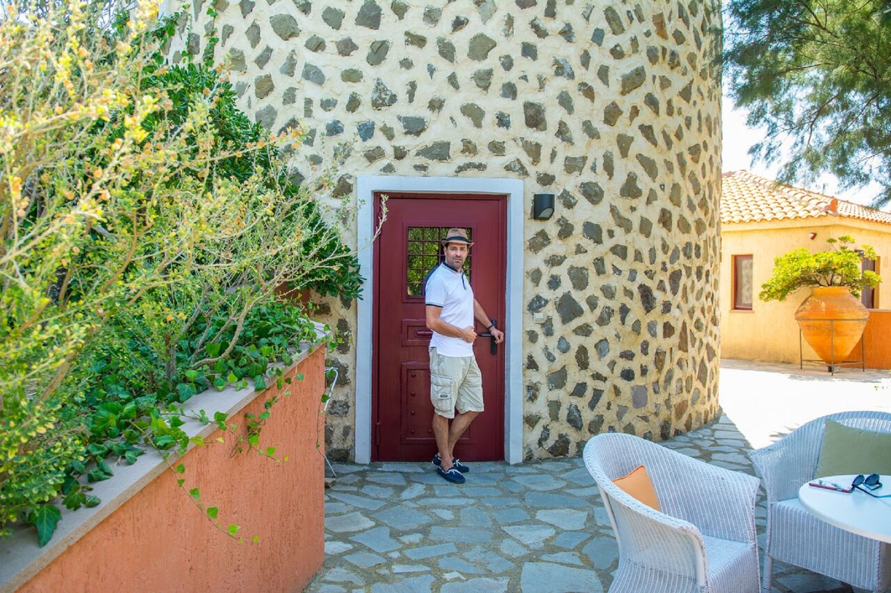 A man standing at a red door of a stone building with a garden and outdoor seating area visible.
