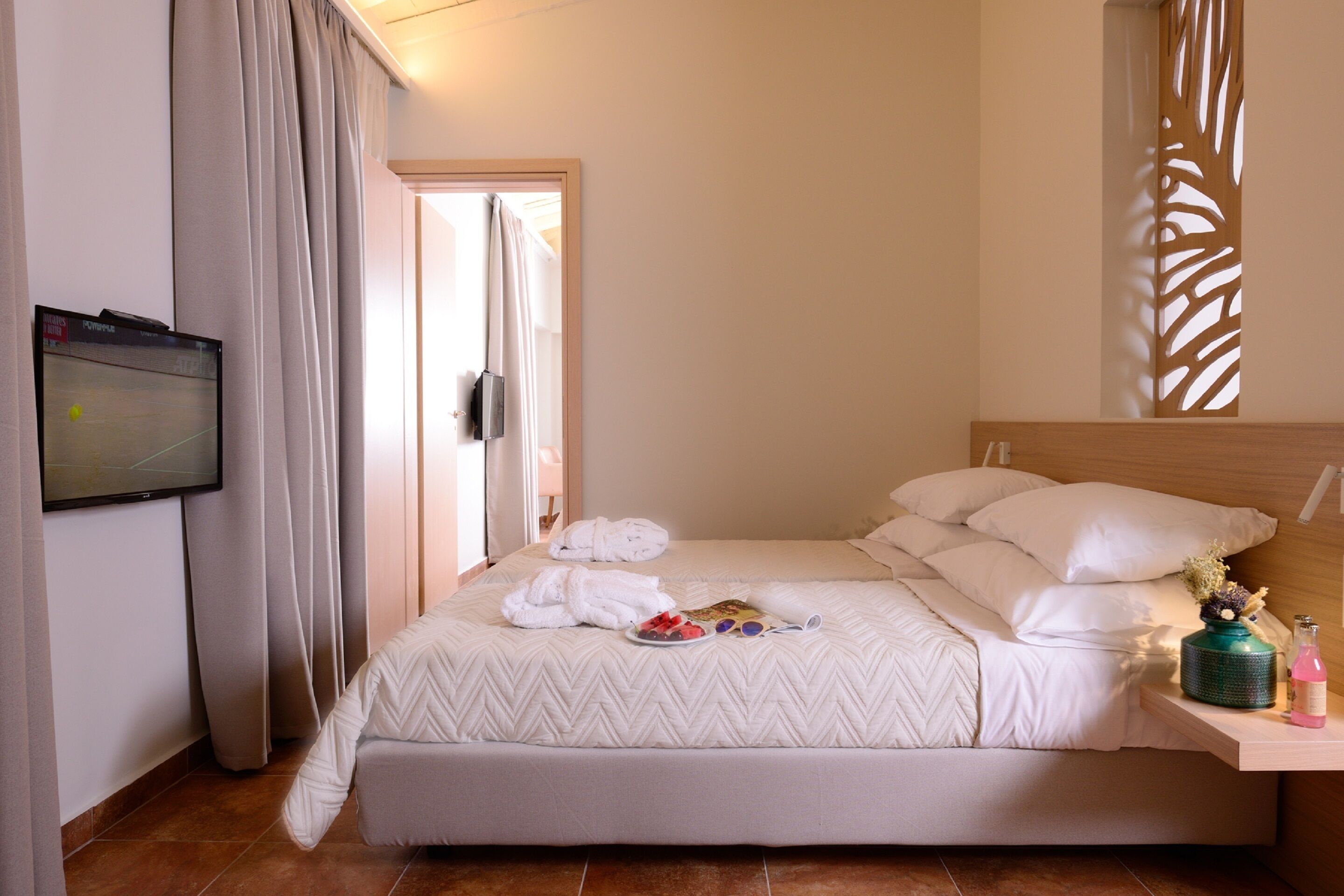 Habitación de hotel con cama, televisión montada en la pared, ventana con cortinas y una mesita con una botella y un florero.