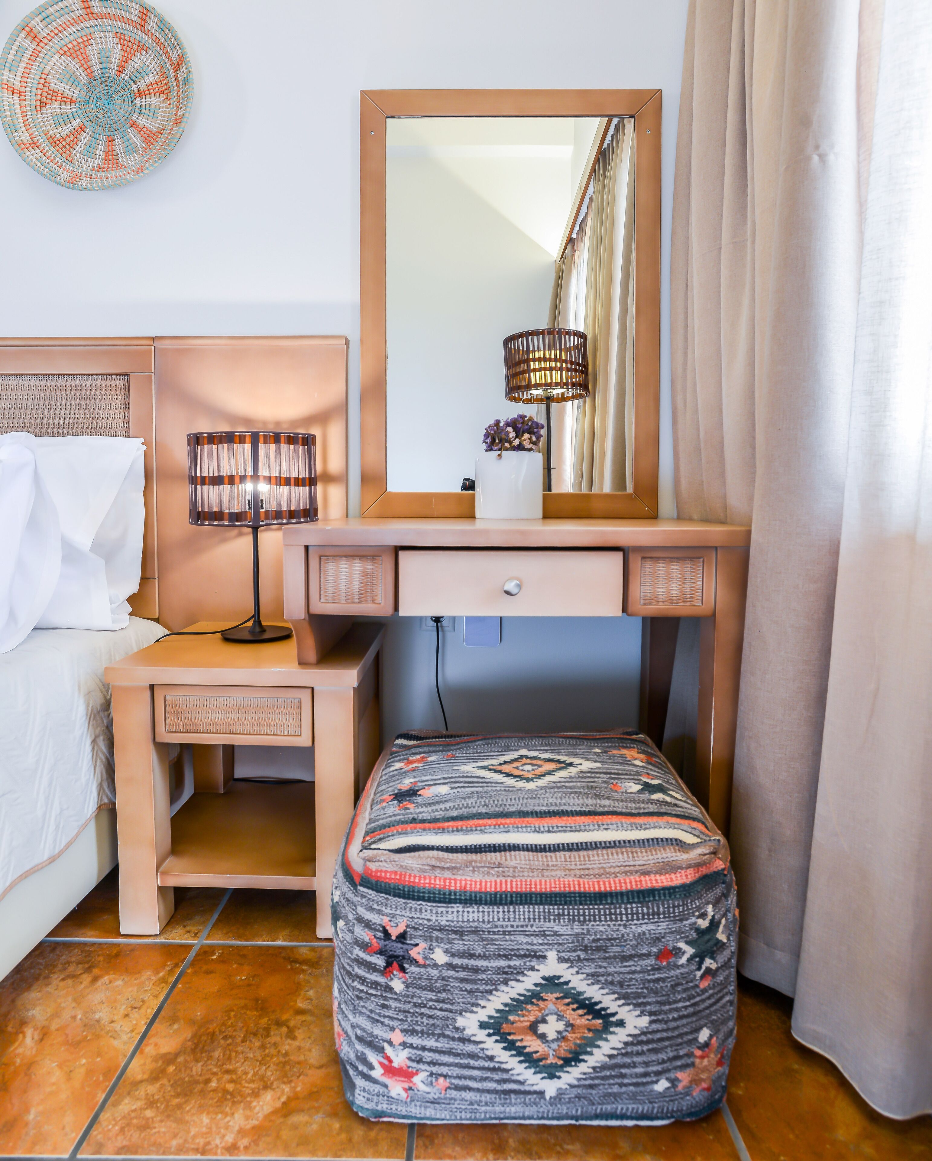 Une chambre à coucher bien rangée, avec un lit, une table de chevet dotée d’une lampe, une commode avec un miroir et un pouf à motifs.