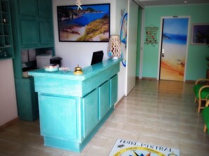 A hotel reception area with a teal reception desk, a painting of a coastal landscape, and a sign indicating the hotel name. - Mistral Hotel (Campo nell'Elba)