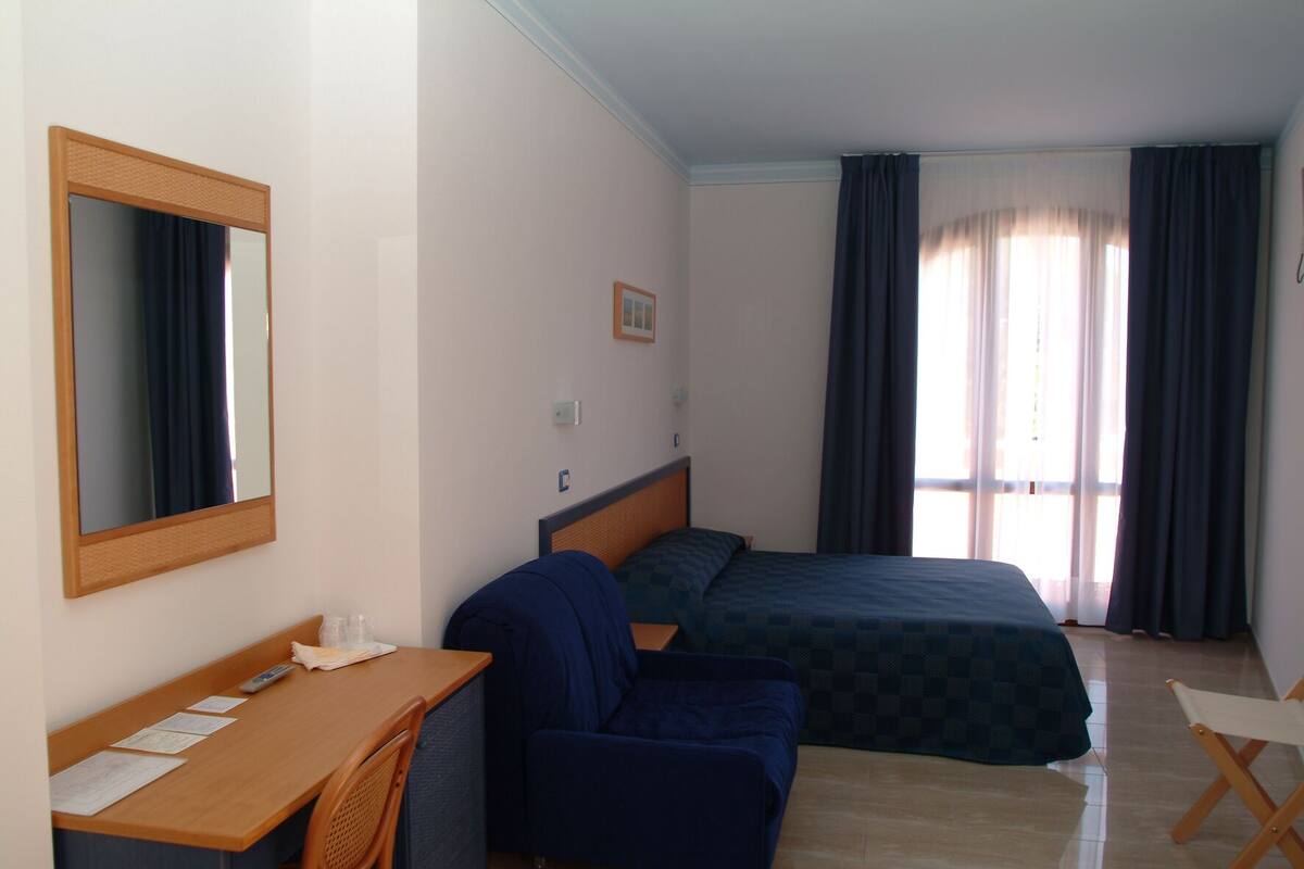 Habitación de hotel con cama, escritorio, silla, espejo y ventana con cortinas.