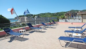 Terraza en la azotea con tumbonas y mesas, banderas, y vistas al mar y las colinas.