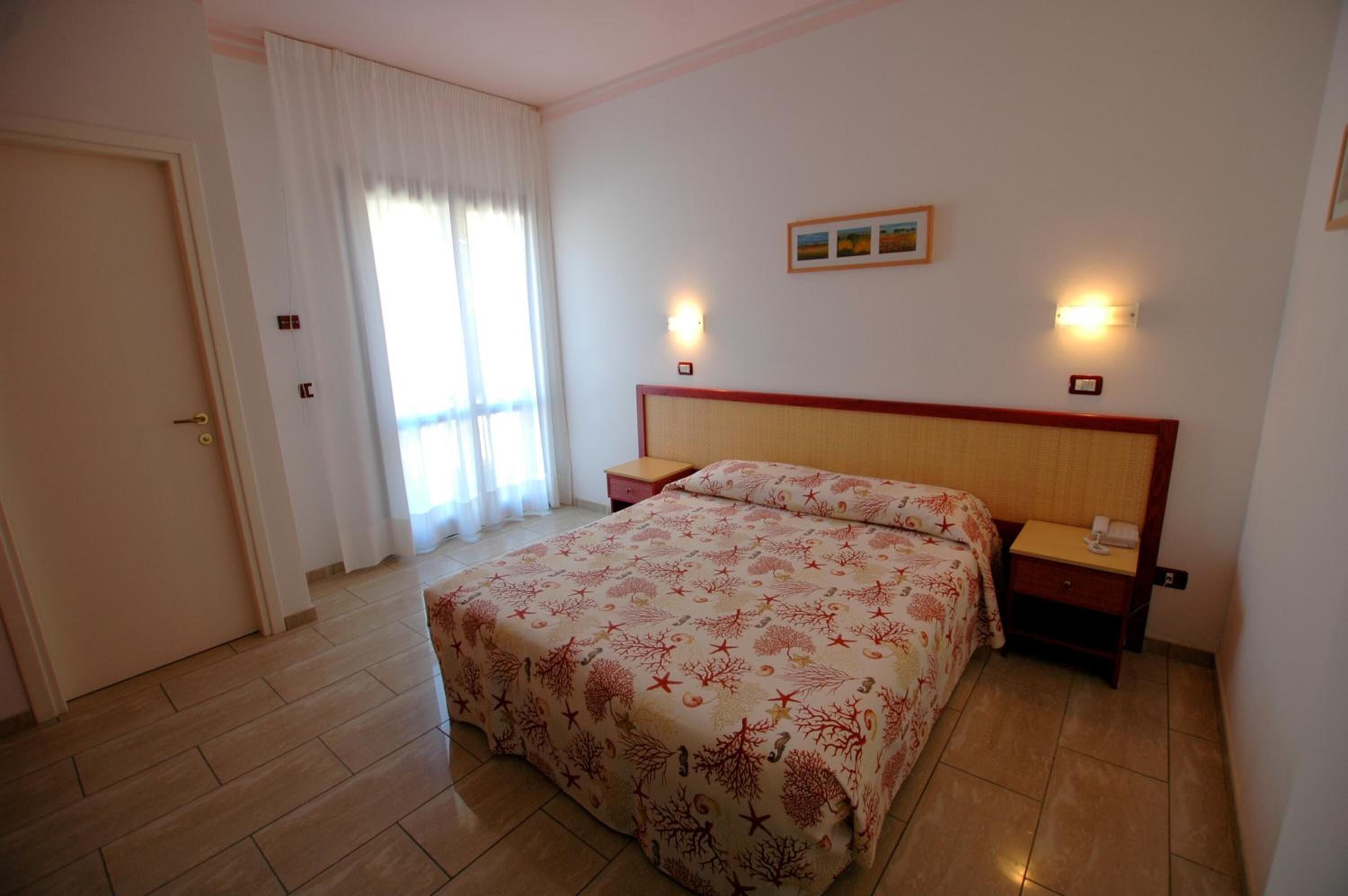 Una habitación de hotel con cama, dos mesitas de noche, una lámpara de pared y cortinas en la ventana.