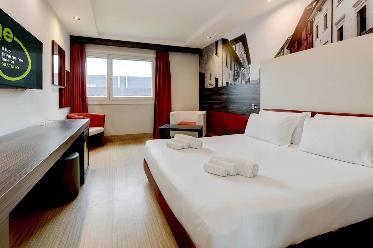 A hotel room with a bed, a desk, a chair, a TV, and a window with curtains.