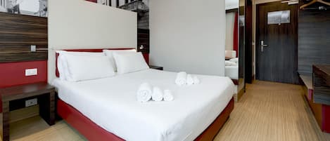 A modern hotel room with a large bed, a wooden nightstand, a red accent wall, and a view of the city through a large window.