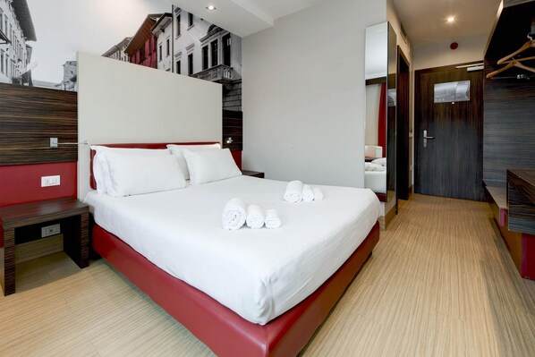 A modern hotel room with a large bed, a wooden nightstand, a red accent wall, and a view of the city through a large window.