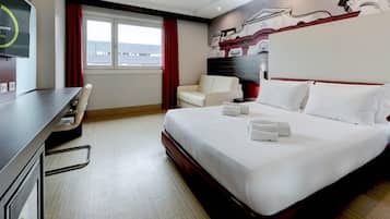 A hotel room with a bed, a desk, a chair, a window with red curtains, and a wall mural.