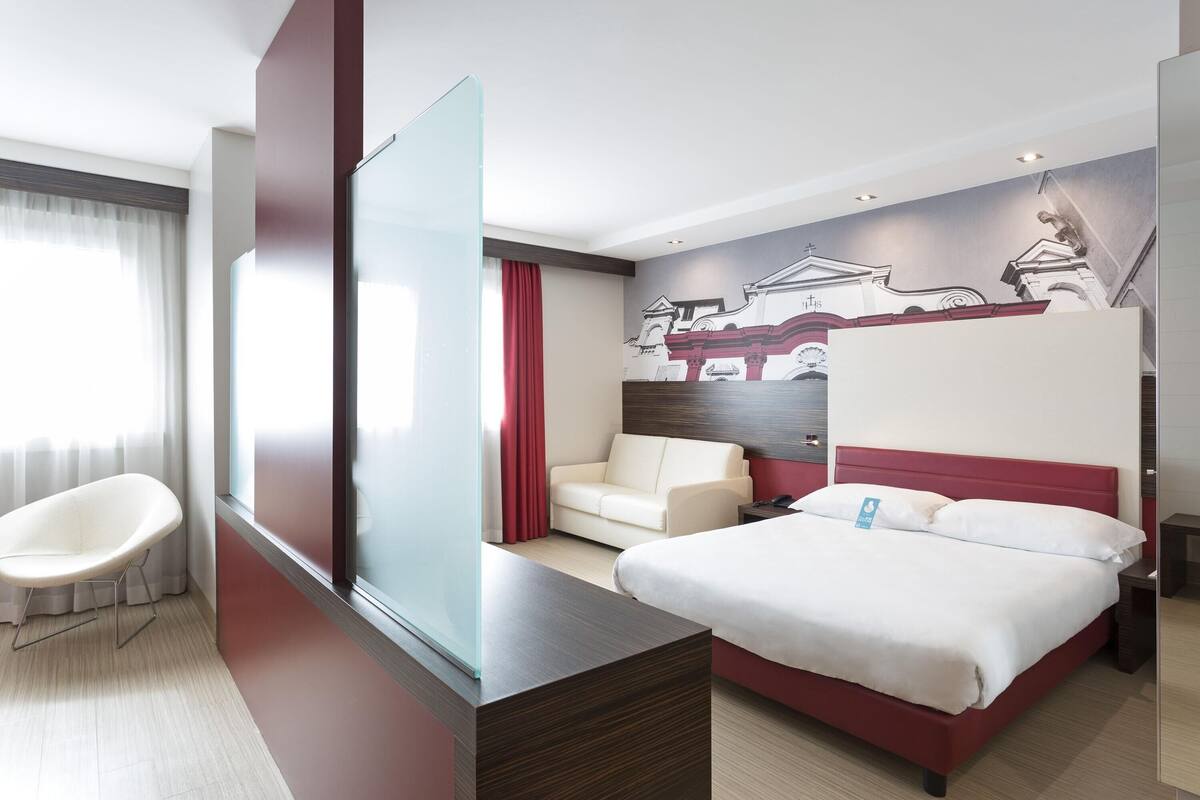 A hotel room with a bed, a sofa, a chair, and a large window.