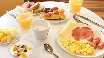 A breakfast plate with toast, croissants, ham, cheese, sausages, scrambled eggs, fruit, and juice.