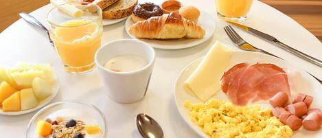 Een ontbijt bord met toast, croissants, ham, kaas, worstjes, roerei, fruit en sap.