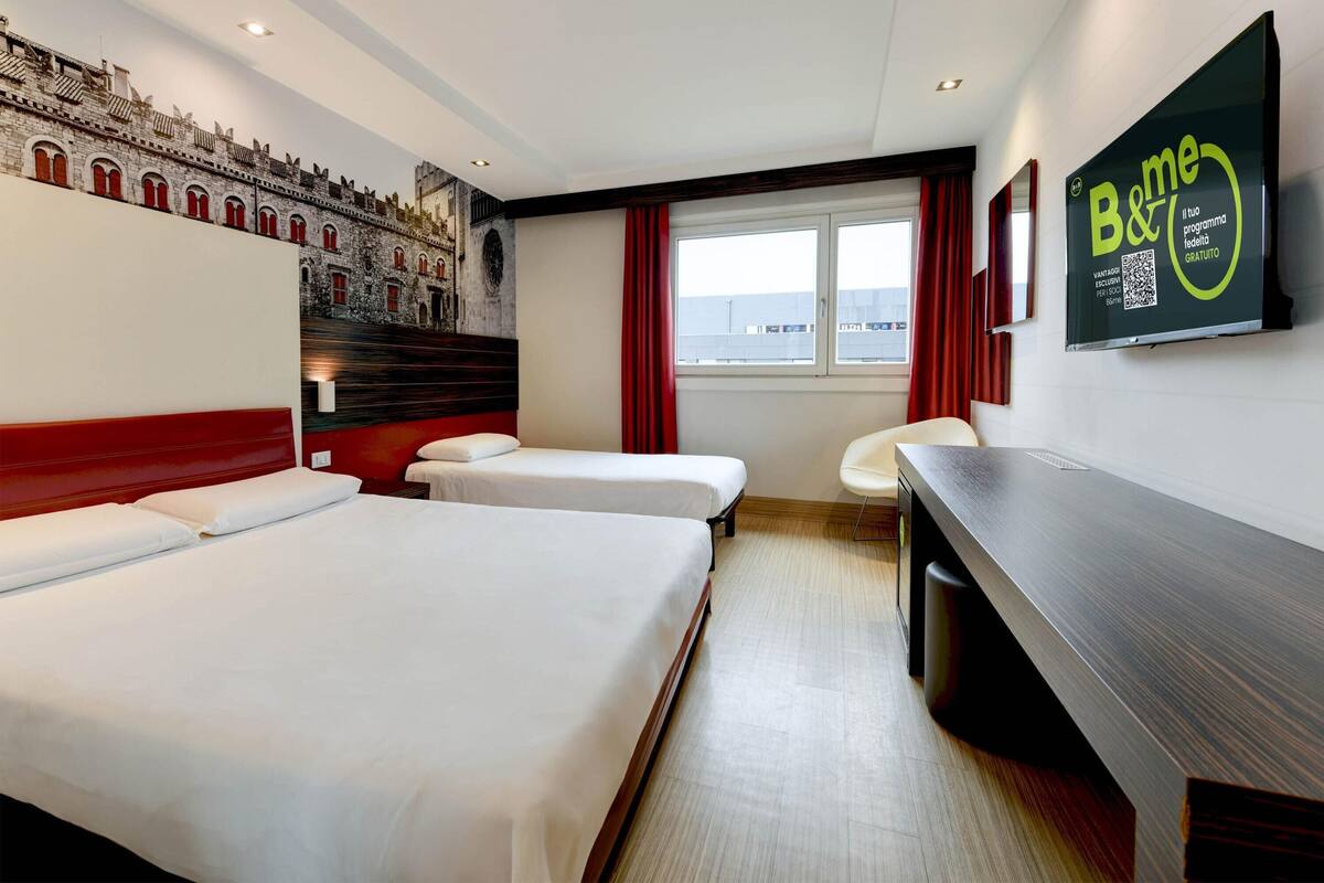 A hotel room with two beds, a desk, a TV, and a mural of a building.