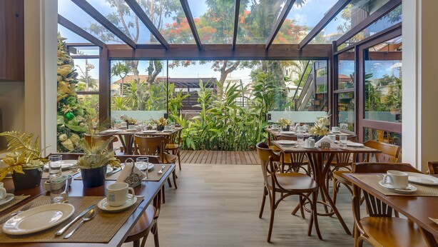 A restaurant with tables set for dining, a view of the garden through large windows, and a wooden ceiling structure. - Nova Pousada dos Chás (Florianópolis)